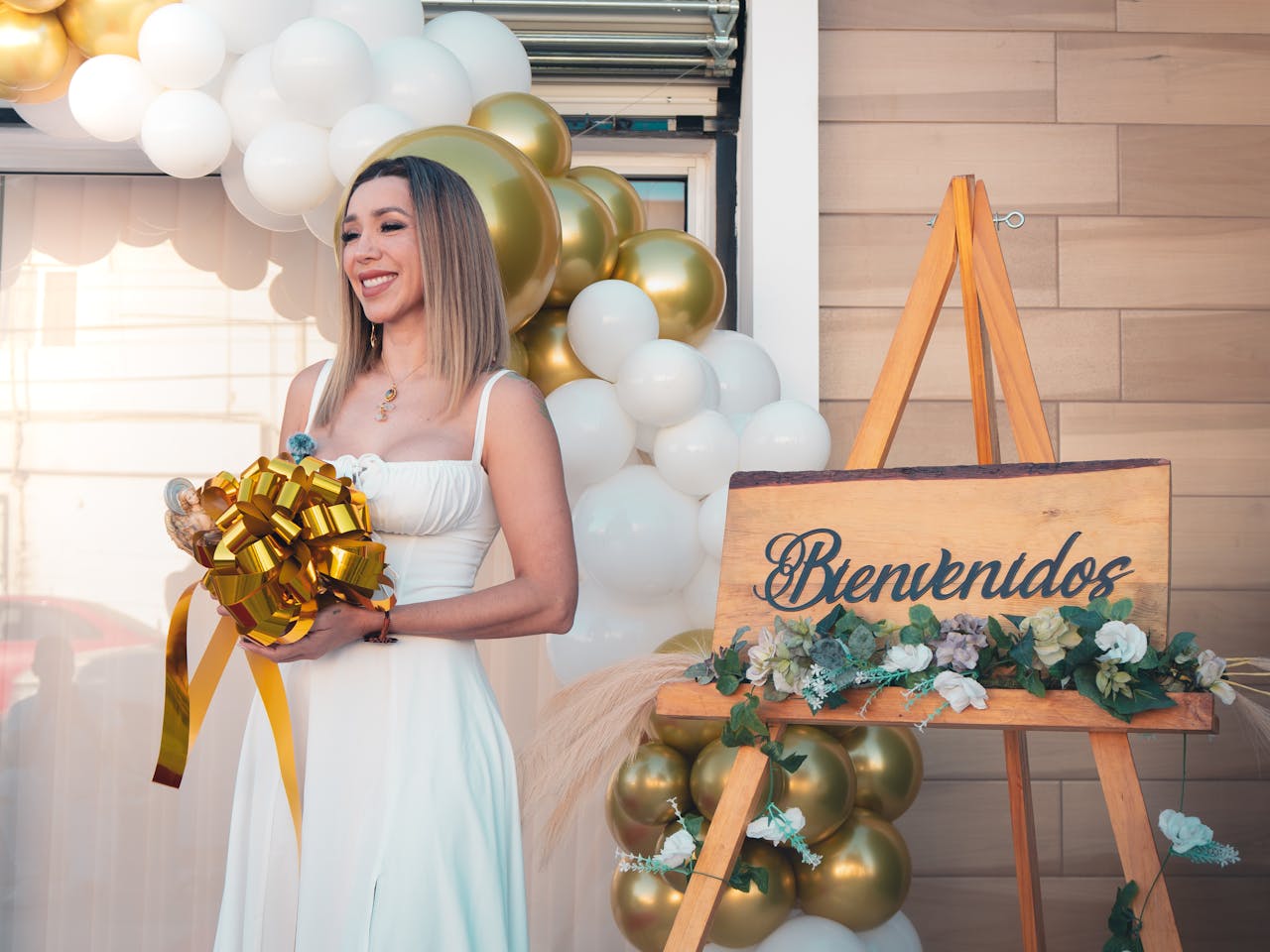 gallery-1 Woman in white holding gold bow at an elegant event with 'Bienvenidos' sign.