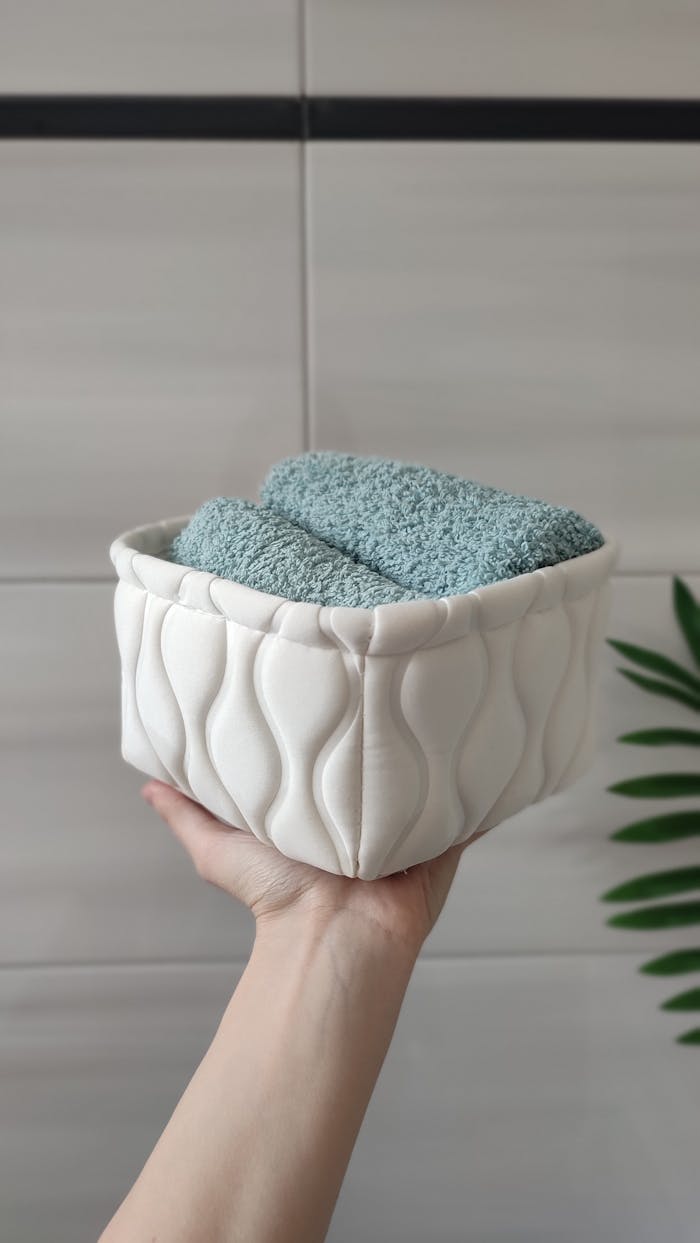 gallery-6 A hand holding a white quilted basket with blue towels against a tiled bathroom wall.