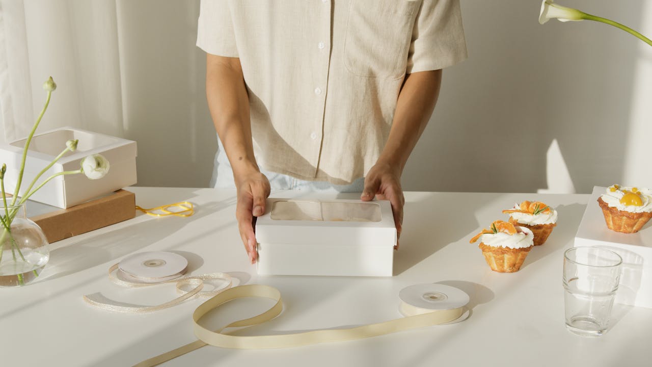 gallery-3 Person packaging cupcakes in a white box surrounded by ribbons and decor on a sunny table.