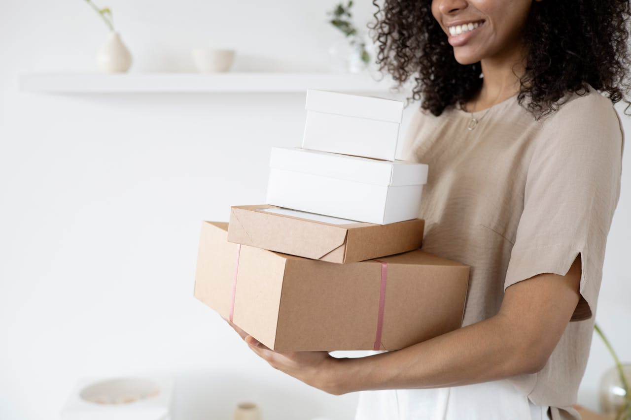 gallery-2 Woman with curly hair carrying stack of packaging boxes in a bright room.