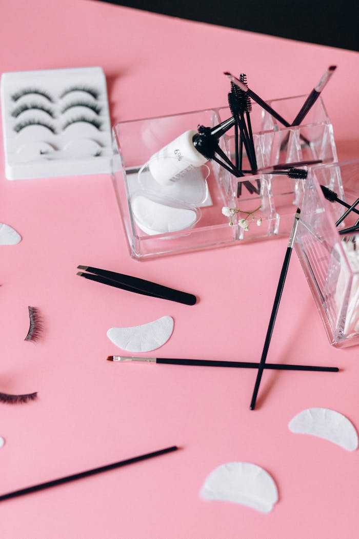 about-01 A detailed flat lay of makeup accessories on a pink background featuring false eyelashes and cosmetic brushes.